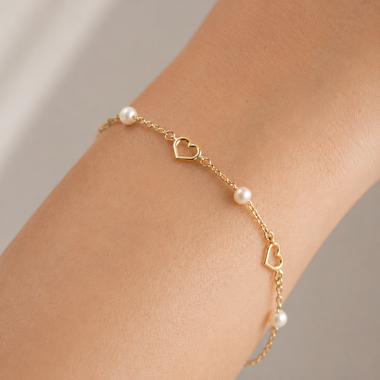 Gold bracelet with heart charms and pearls on a wrist against a neutral background
