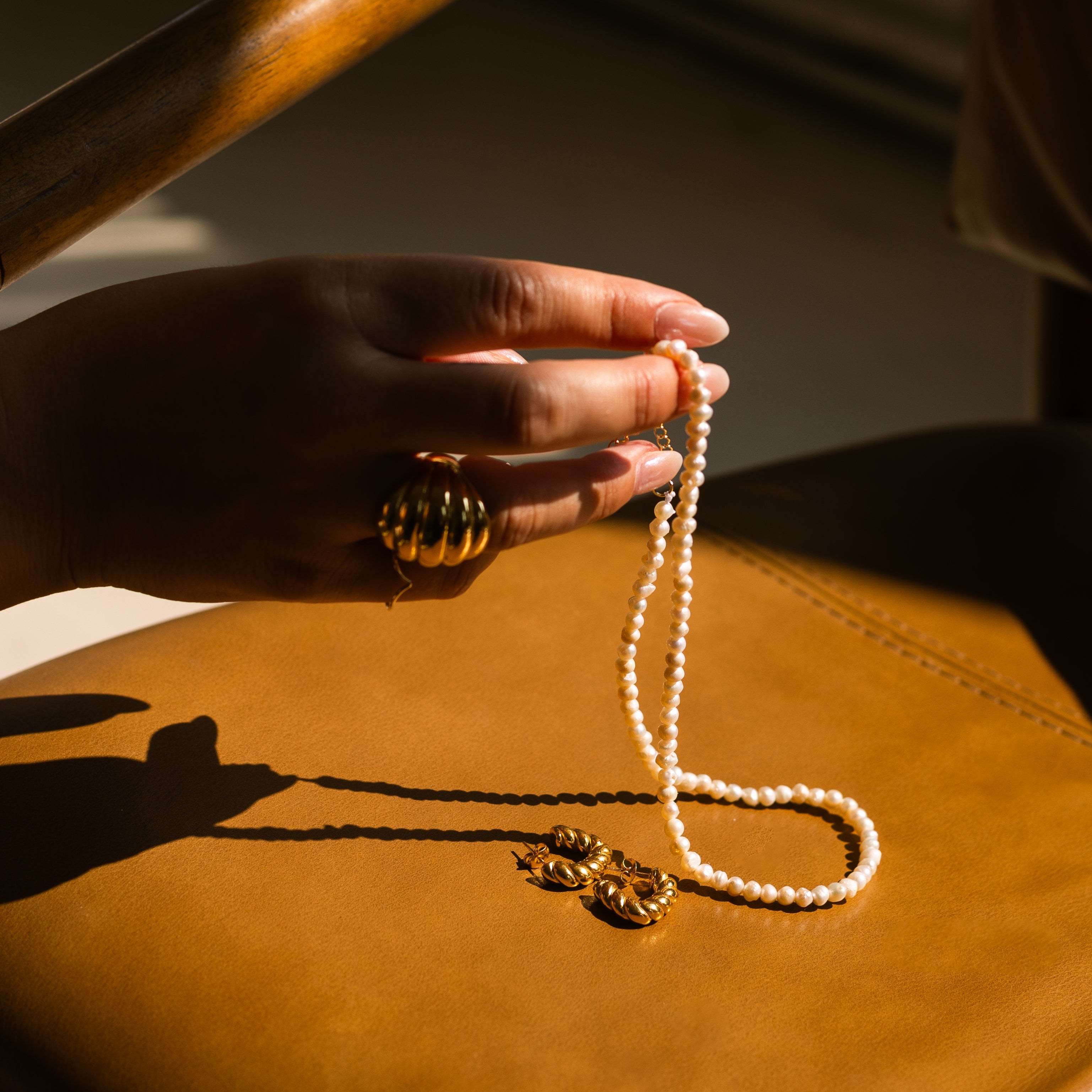 A close-up of a hand holding a delicate pearl necklace with gold-toned charms, resting on a tan leather surface with dramatic sunlight and shadows.
