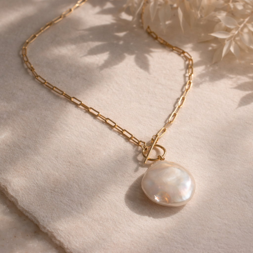 Gold paperclip necklace with a baroque pearl pendant on a textured surface with leaf shadows