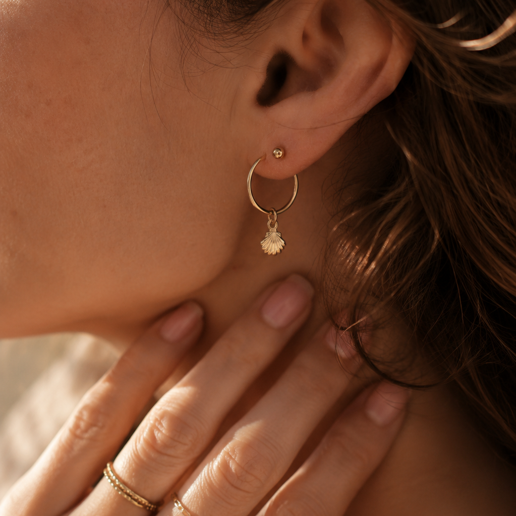 Close-up of a person wearing gold hoop earrings and rings on a neutral background