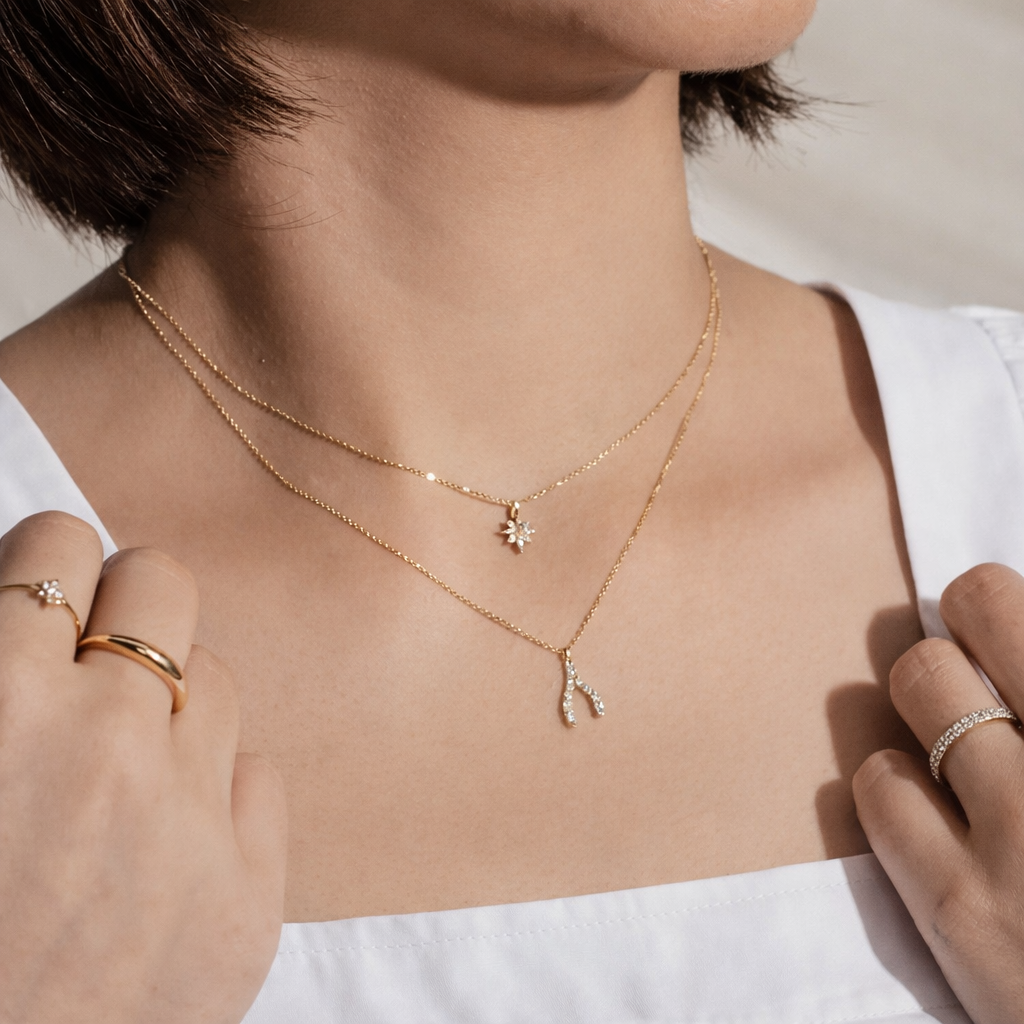 Close-up of a person wearing two gold necklaces with wishbone pendant on a neutral background.